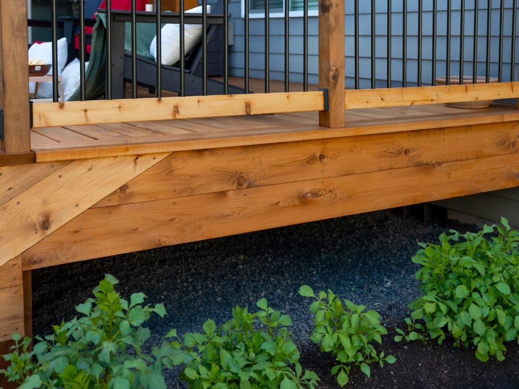 A close-up view of a wooden deck with stairs, featuring natural wood grain and metal railings. Green leafy plants grow in the soil beneath the deck, and part of a house with gray siding is visible in the background.