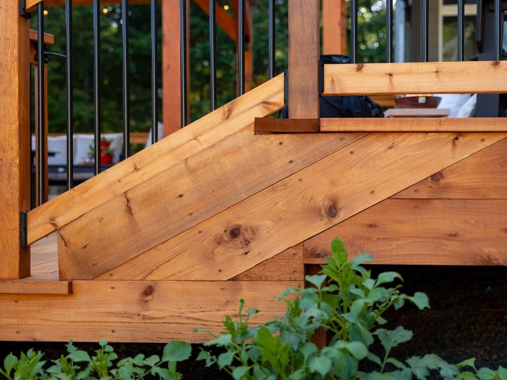 Close-up of a wooden outdoor staircase with metal railings, surrounded by green plants at the base and trees blurred in the background.