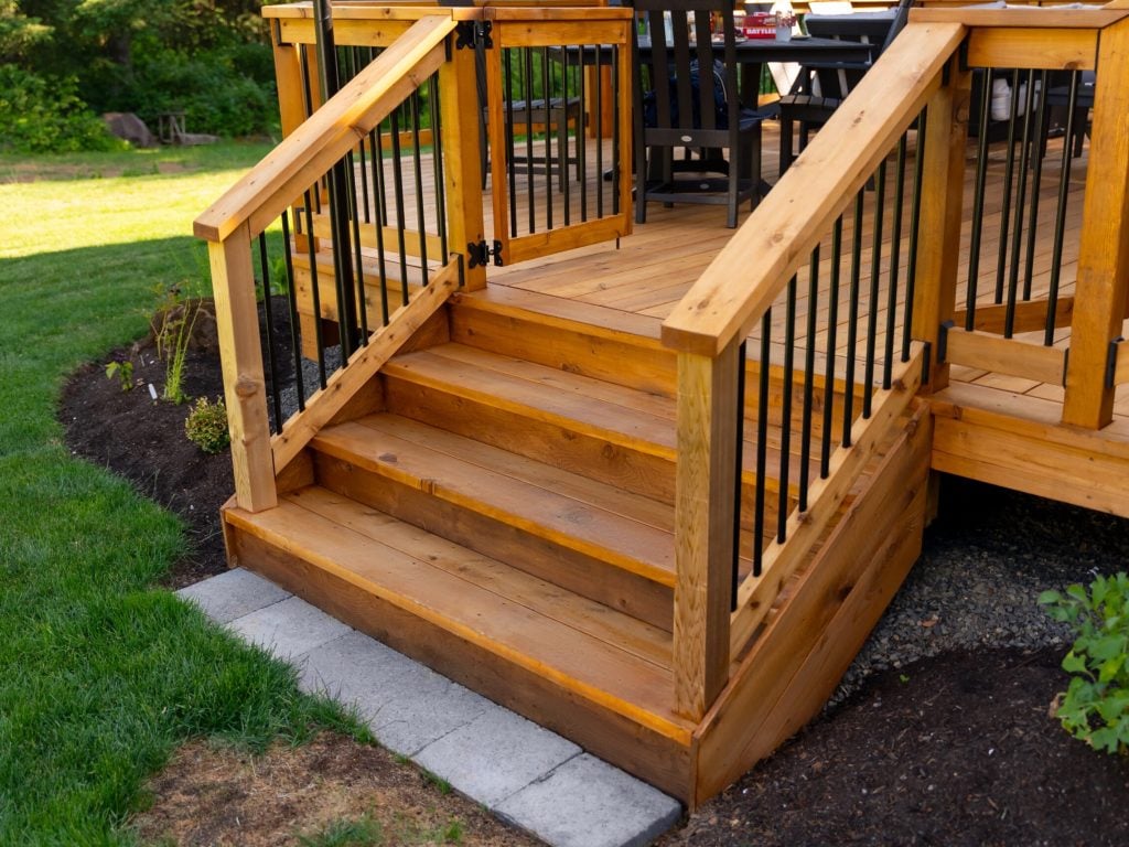 Wooden deck steps with black metal railings lead from a raised deck to a grassy yard, bordered by a garden bed with mulch and plants. The deck features outdoor seating in the background.