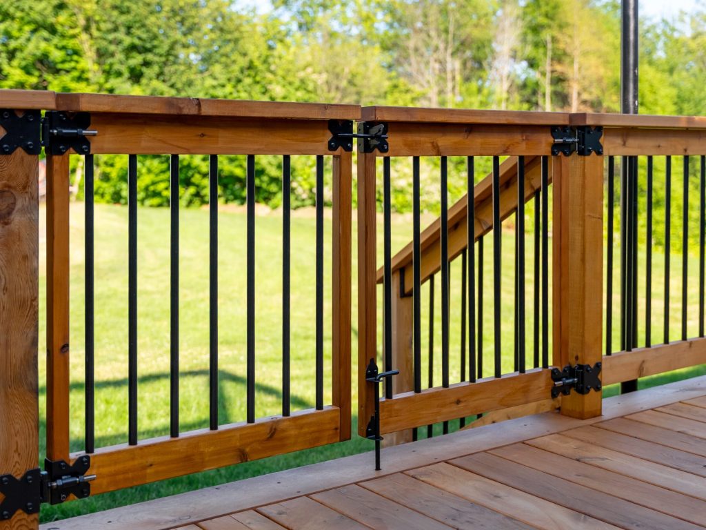A wooden deck with a safety gate and black metal railings, overlooking a green, grassy yard with trees in the background on a sunny day.