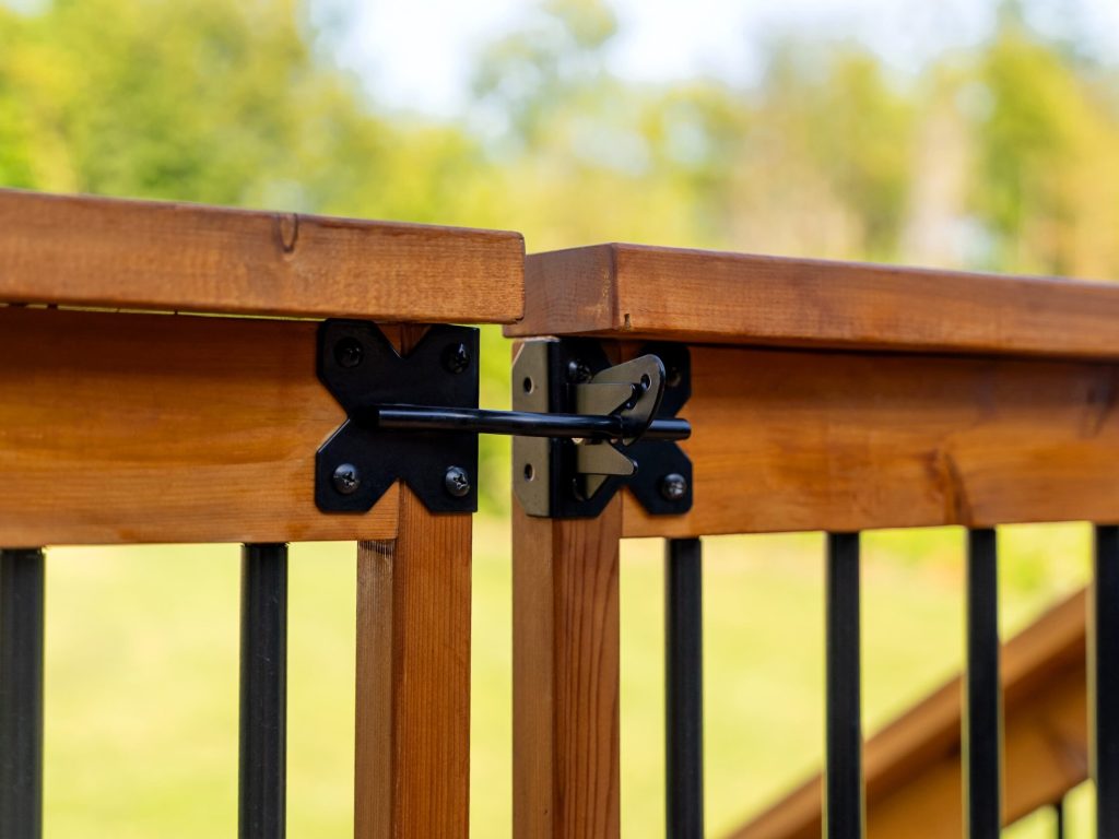 Close-up of a wooden gate with black metal bars and a black metal latch securing the two sections. The background is blurred greenery, suggesting an outdoor setting.