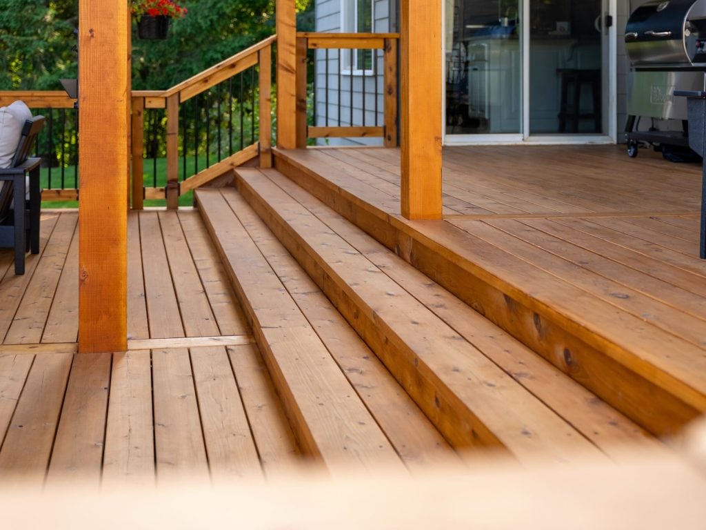 A close-up view of wooden deck stairs leading up to a patio area with railings and outdoor furniture; a grill and sliding glass door are visible in the background.