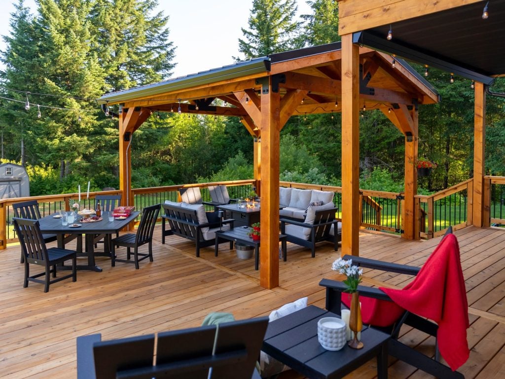 A spacious wooden deck with a covered pergola, outdoor seating, a dining table set for a meal, black chairs, and lush green trees in the background on a sunny day.