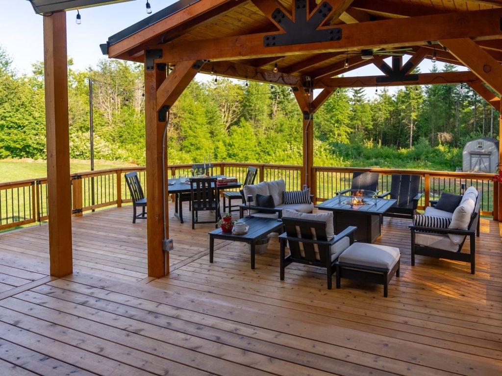 A spacious wooden deck with a covered pergola, outdoor seating, tables, and a fire pit, surrounded by green trees and a grassy yard on a sunny day.