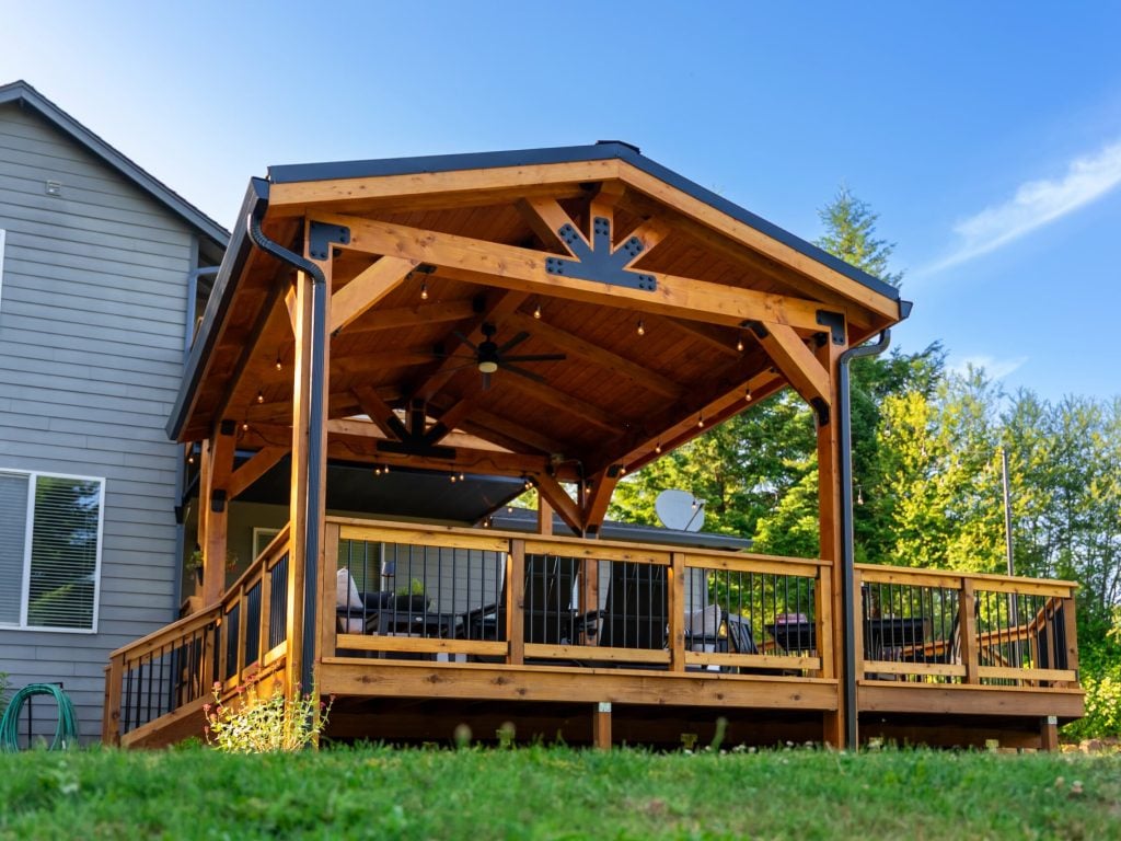A modern house with a spacious wooden deck and a covered patio, featuring black railings and outdoor furniture, surrounded by green trees and grass under a clear blue sky.