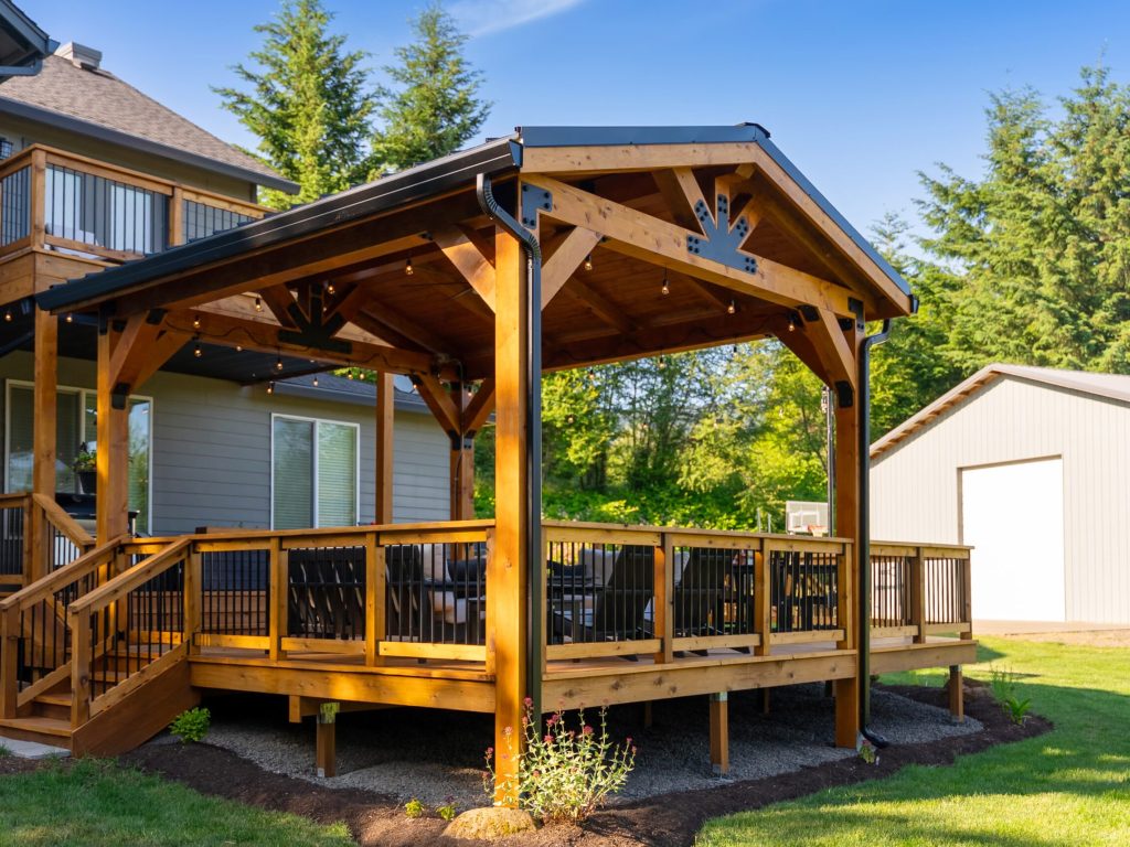 A wooden covered deck with black railings attached to a house, featuring outdoor seating and string lights, surrounded by greenery and a large shed in the background.