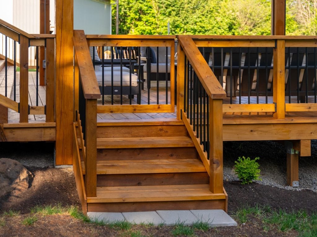 Wooden steps with black metal railings lead up to a wooden deck surrounded by greenery and patio furniture. Sunlight highlights the warm tones of the wood.