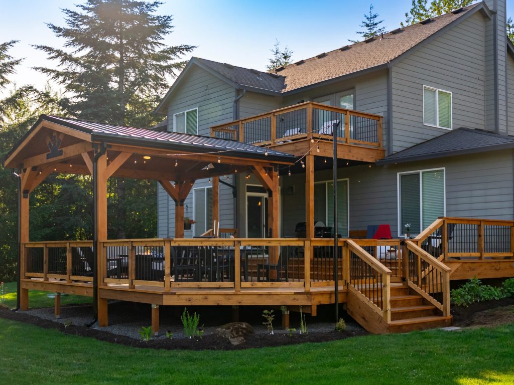 A modern two-story gray house with a large wooden deck, stairs, covered seating area, string lights, and railing, surrounded by green grass and trees on a sunny day.
