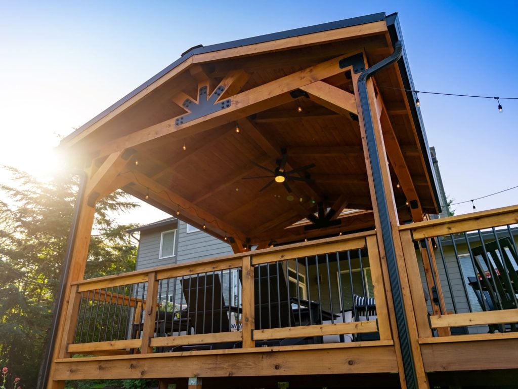 A wooden porch with a peaked roof and string lights is attached to a house. The sun is shining brightly behind the structure, casting a warm glow, and trees are visible in the background.