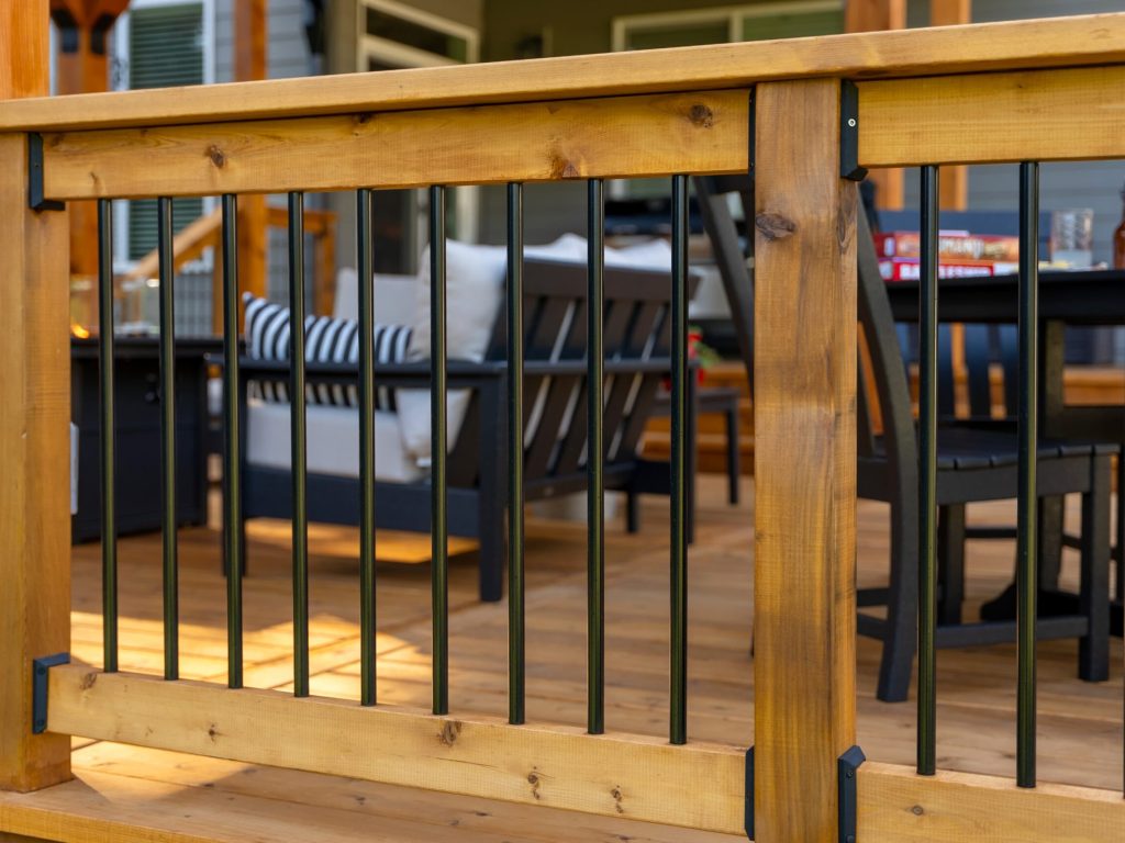Close-up of a wooden deck railing with black metal spindles, showing an outdoor seating area with cushioned chairs, a striped pillow, and a table in the background. The deck appears to be part of a porch or patio.