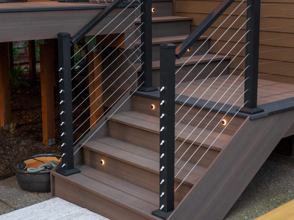 Modern outdoor wooden stairs with cable railing and built-in step lights lead up to a deck attached to a brown house. A gray mat and a metal dog bowl sit at the bottom of the stairs.