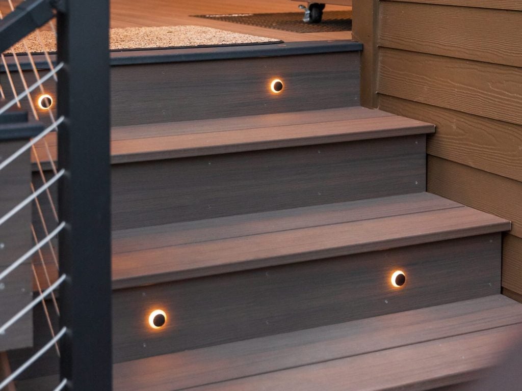Three outdoor wooden steps with built-in circular lights on each riser, leading to a deck with a metal cable railing; a brown wall and lush greenery are visible in the background.