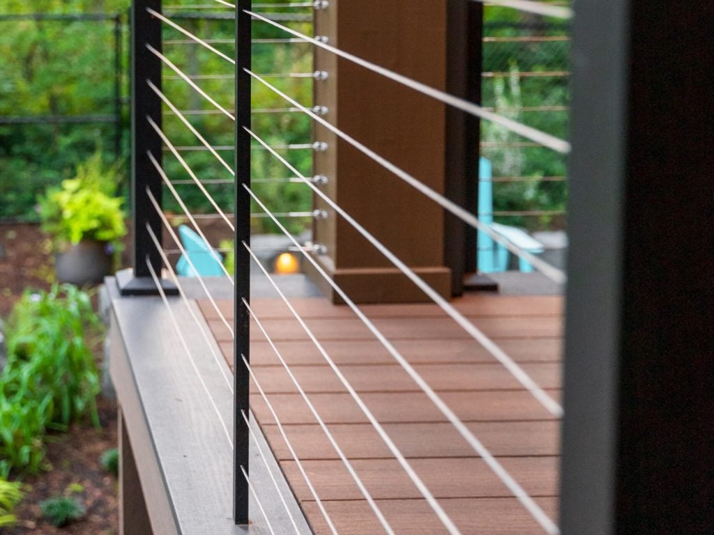 Close-up view of a modern deck with dark wood flooring and sleek cable railing, overlooking a green, landscaped backyard with trees and plants in the background.