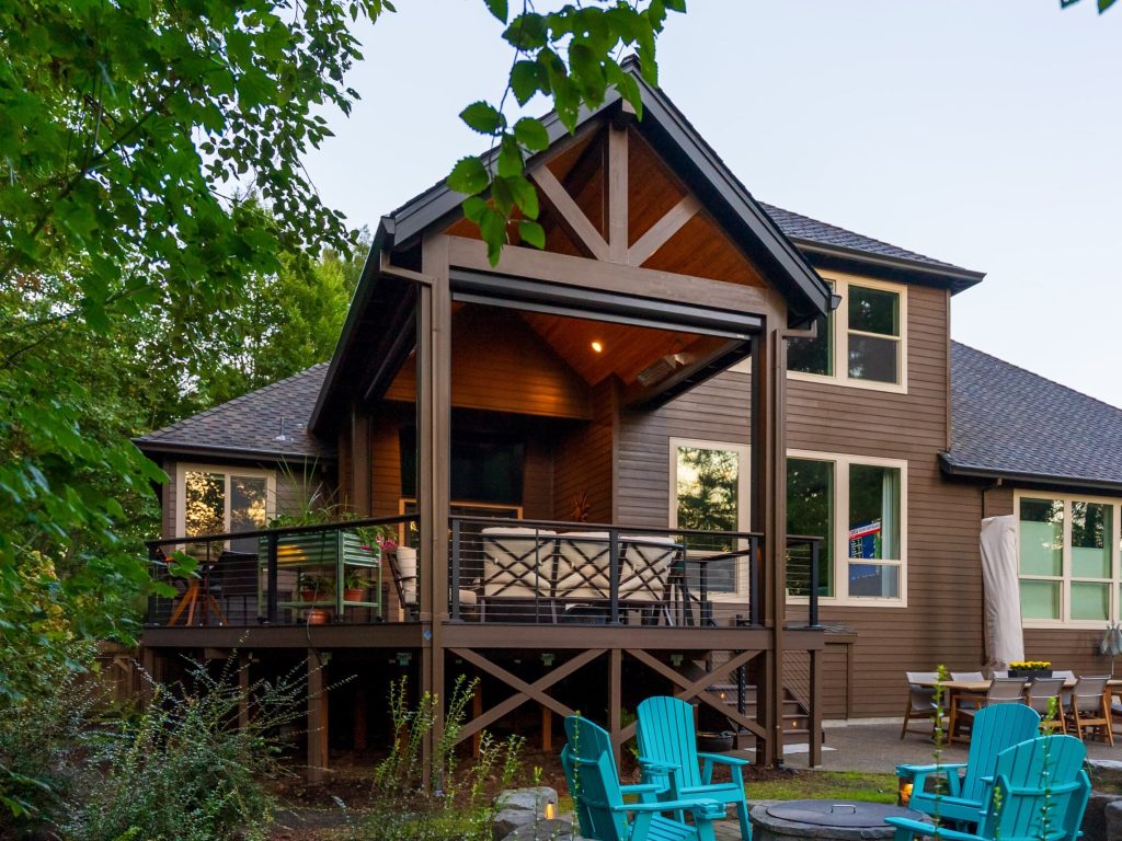 A modern two-story house with a covered elevated deck, surrounded by trees. The deck has outdoor seating and a railing. Bright blue Adirondack chairs and a fire pit are arranged in the backyard.