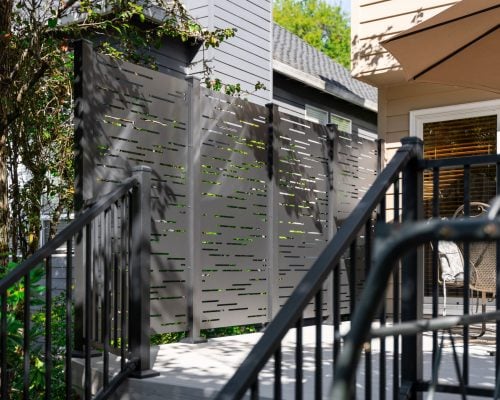 A modern patio featuring a decorative privacy screen with horizontal cutouts, surrounded by greenery, black railings, and outdoor furniture under a large umbrella.