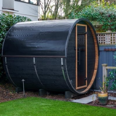 A barrel-shaped black sauna with a wooden door sits in a garden, surrounded by green bushes and a manicured lawn, next to a modern house with blue siding.
