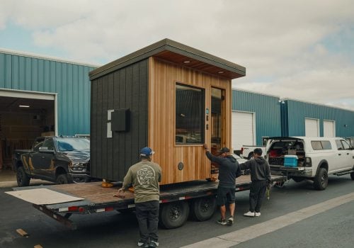 Three people load a small modern wooden tiny house onto a flatbed trailer, parked outside blue industrial buildings under a cloudy sky.