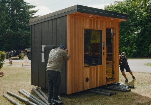 Two people push a small modern wooden shed on pipes across a grassy yard, using the pipes as rollers to move the building. Trees and a driveway are visible in the background.