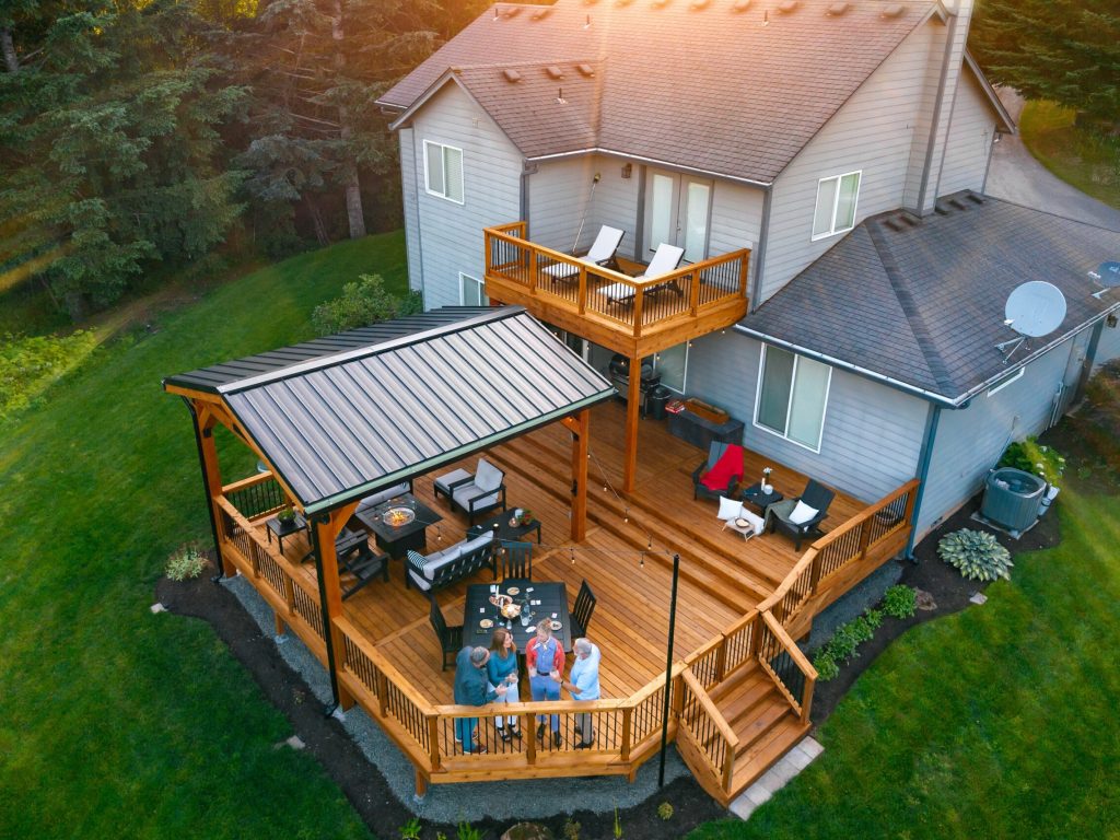 Aerial view of a modern house with a large, multi-level wooden deck. Four people are gathered around a table with food, and lounge chairs are arranged on the deck. The home is surrounded by green lawn and trees.