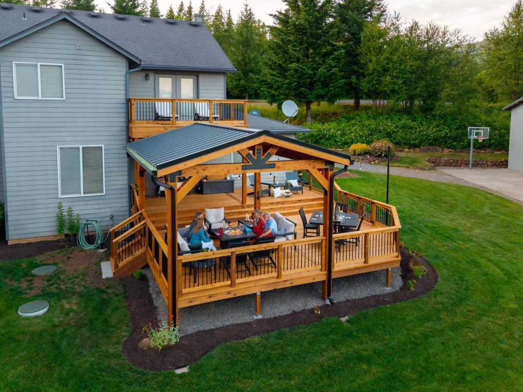 A group of people sit around a table on a wooden deck attached to a gray house, surrounded by green grass and trees. The deck has a covered section, railings, and outdoor furniture.
