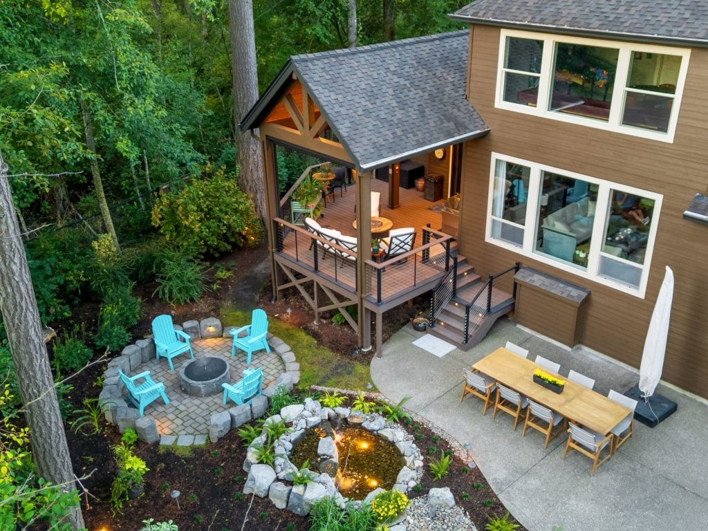A backyard features a raised wooden deck with seating, a dining area on a concrete patio, a circular fire pit with blue chairs, and a small landscaped pond, surrounded by trees and greenery.