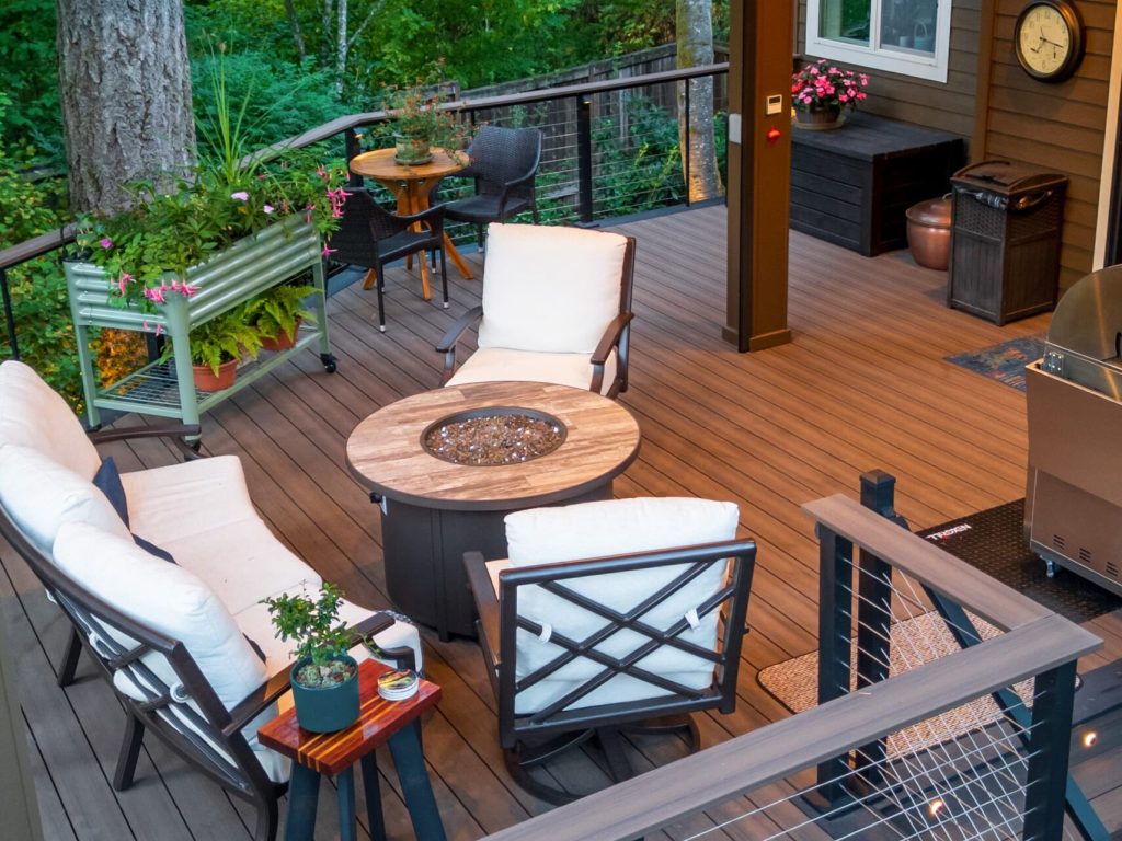A cozy outdoor deck with cushioned chairs around a fire pit table, potted plants, a grill, and a clock on the house wall, surrounded by trees and greenery.