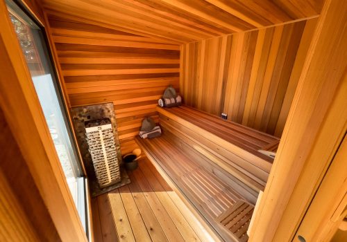A wooden sauna interior with tiered benches, a bucket and ladle on the floor, a heater with rocks, and neatly rolled towels placed on the benches. Sunlight streams in through a window, creating a warm atmosphere.