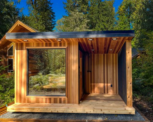 A modern sauna with large windows and vertical wood siding stands in a wooded area, surrounded by tall trees. The house features a covered porch and a flat roof with recessed lights.