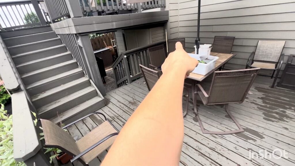 A persons arm points towards a patio table with chairs and a closed umbrella on a weathered wooden deck, next to stairs leading to an upper level.
