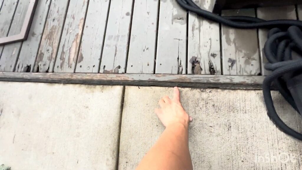 A persons arm points at the edge where a weathered wooden deck meets a concrete surface; a black hose is coiled nearby.