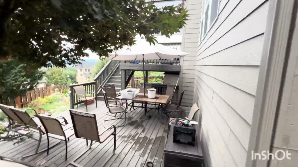 A wooden deck with patio chairs, a table, a large umbrella, and a staircase leading up. There are trees overhead and a partial view of buildings and greenery in the background.