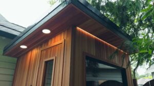 A wood-sided backyard structure with a flat roof, featuring recessed lighting and under-eave LED accent lights.