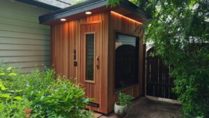 A cedar sauna with a glass door and window installed in a backyard next to a house and green plants.
