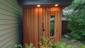 A backyard cedar sauna featuring a door with a vertical window, exterior lighting, and control panels.