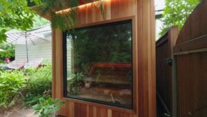 Exterior view of a cedar-paneled sauna featuring a large glass window that reveals interior wooden benches and an illuminated red heating element.