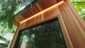 Exterior corner of a wooden building featuring vertical cedar siding, a large glass window, and integrated LED strip lighting under the roof overhang.