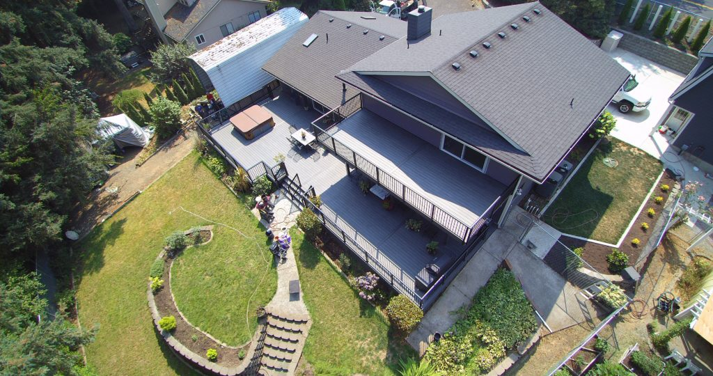 Aerial view of a house with a large deck, hot tub, and fenced yard. There’s a garden with stone pathways, terraced landscaping, and outdoor seating. The home is surrounded by trees and neighboring houses.