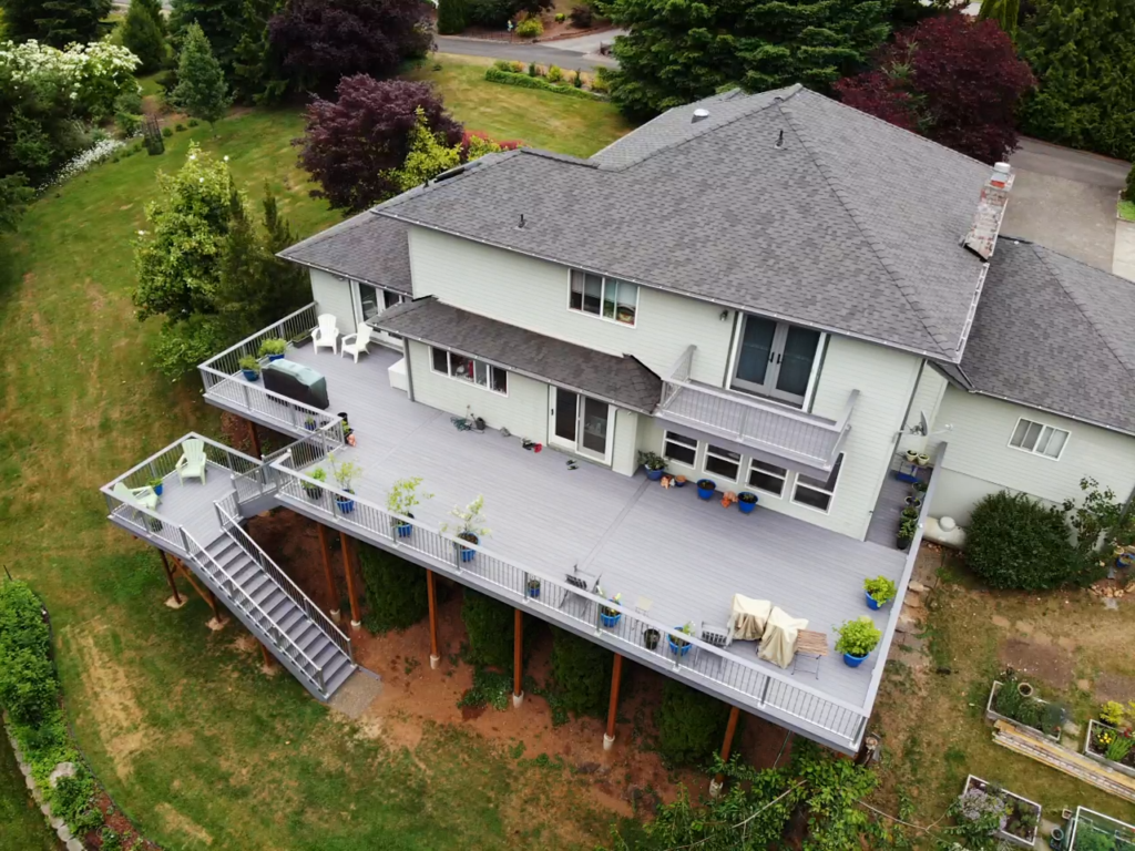 Aerial view of a large gray house with an expansive raised deck featuring potted plants, outdoor furniture, and stairs leading to a grassy yard bordered by trees and a driveway.