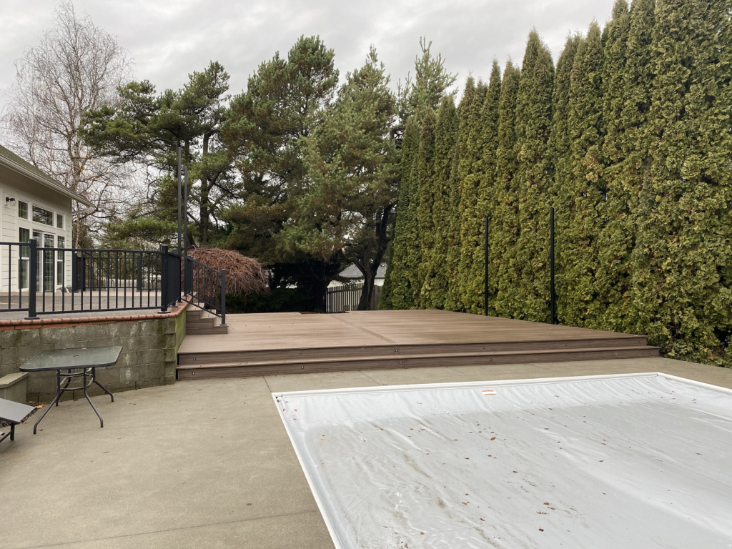 A backyard with a covered pool, concrete patio, glass-top table, raised wooden deck, black railing, and tall evergreen trees along the right side.
