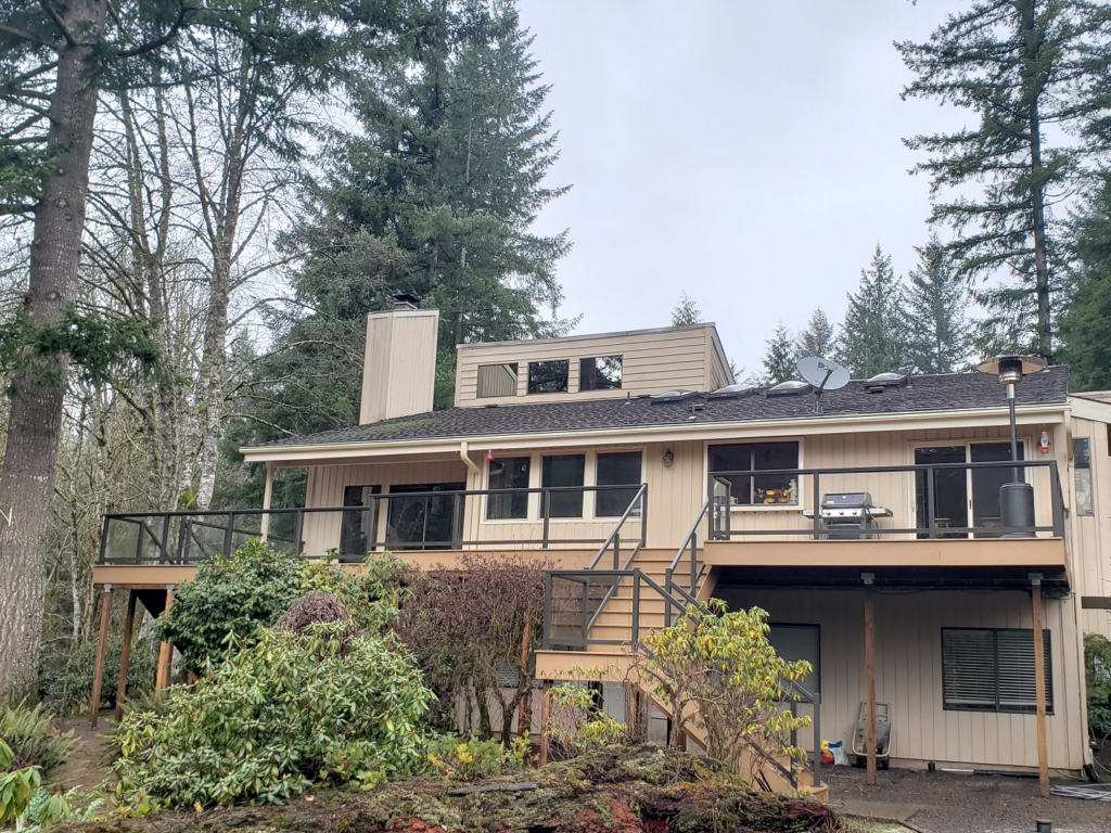 A beige two-story house with large windows, decks with railings, and a staircase, surrounded by trees and shrubs on a cloudy day.