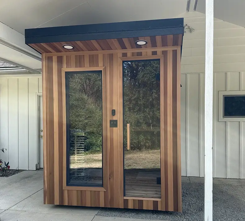 A small modern wooden shed with vertical light and dark wood panels, large glass door and window, flat black roof, and two built-in ceiling lights, standing next to a white house.