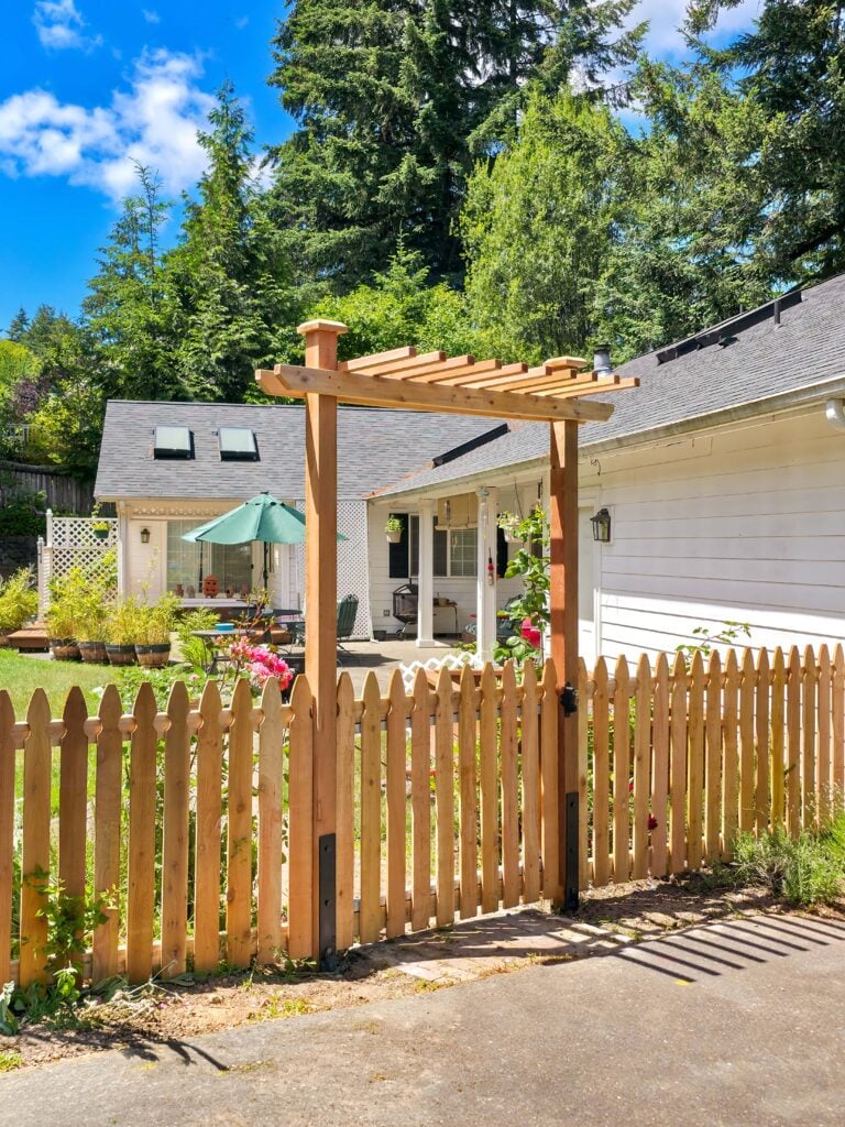A wooden arbor gate in a picket fence opens to a sunny yard with green grass, a patio umbrella, flowers, and a white house, surrounded by tall trees under a blue sky.