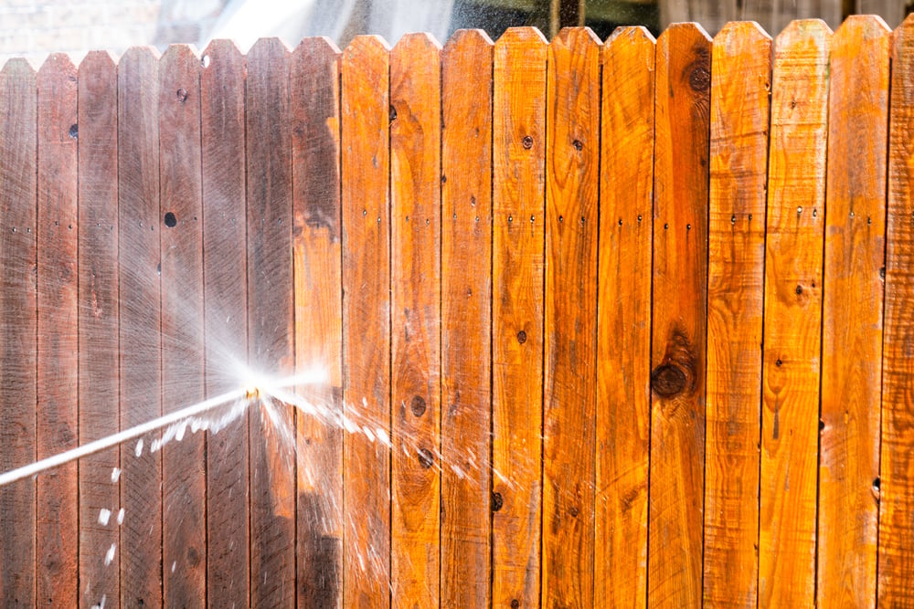A stream of water sprays through a hole in a wooden fence, creating a burst of mist against the bright, sunlit wood.