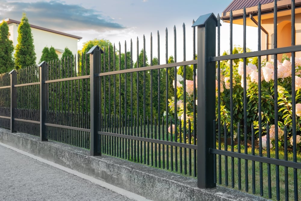 A black metal fence with pointed tops runs along a concrete edge, enclosing a garden with green shrubs and blooming flowers beside a yellow house. The sky is partly cloudy.