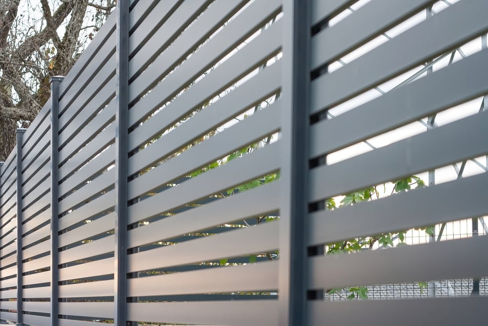Modern gray metal slat fence with horizontal panels, partially revealing trees and greenery behind it. Sunlight filters through the slats, creating a pattern of light and shadow.