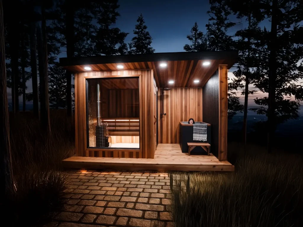 A modern wooden sauna cabin with large glass window sits in a forest at dusk. A person relaxes in a small tub on the deck. Pathway stones lead to the cabin, surrounded by tall grass and silhouetted trees.