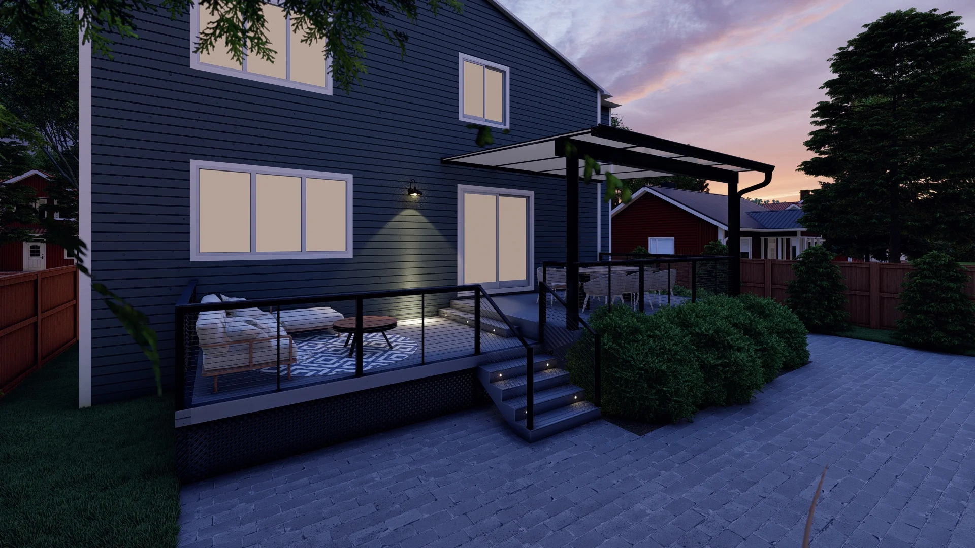 A modern backyard patio at dusk features a raised deck with glass railing, outdoor seating, a rug, illuminated steps, and overhead canopy attached to a dark blue house. Bushes line the deck and pavers cover the ground below.
