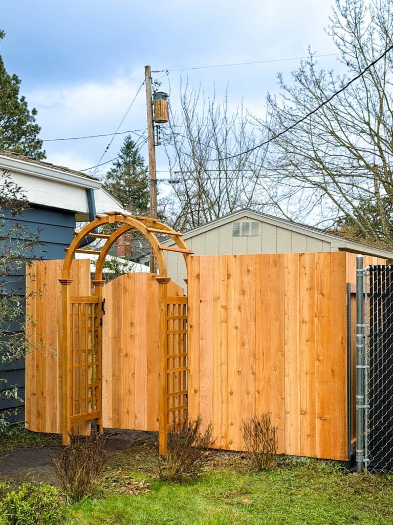 A wooden garden gate with an arched trellis stands in a new wooden fence beside a blue house, with a shed, trees, power lines, and a cloudy sky in the background. Grass and shrubs are in the foreground.